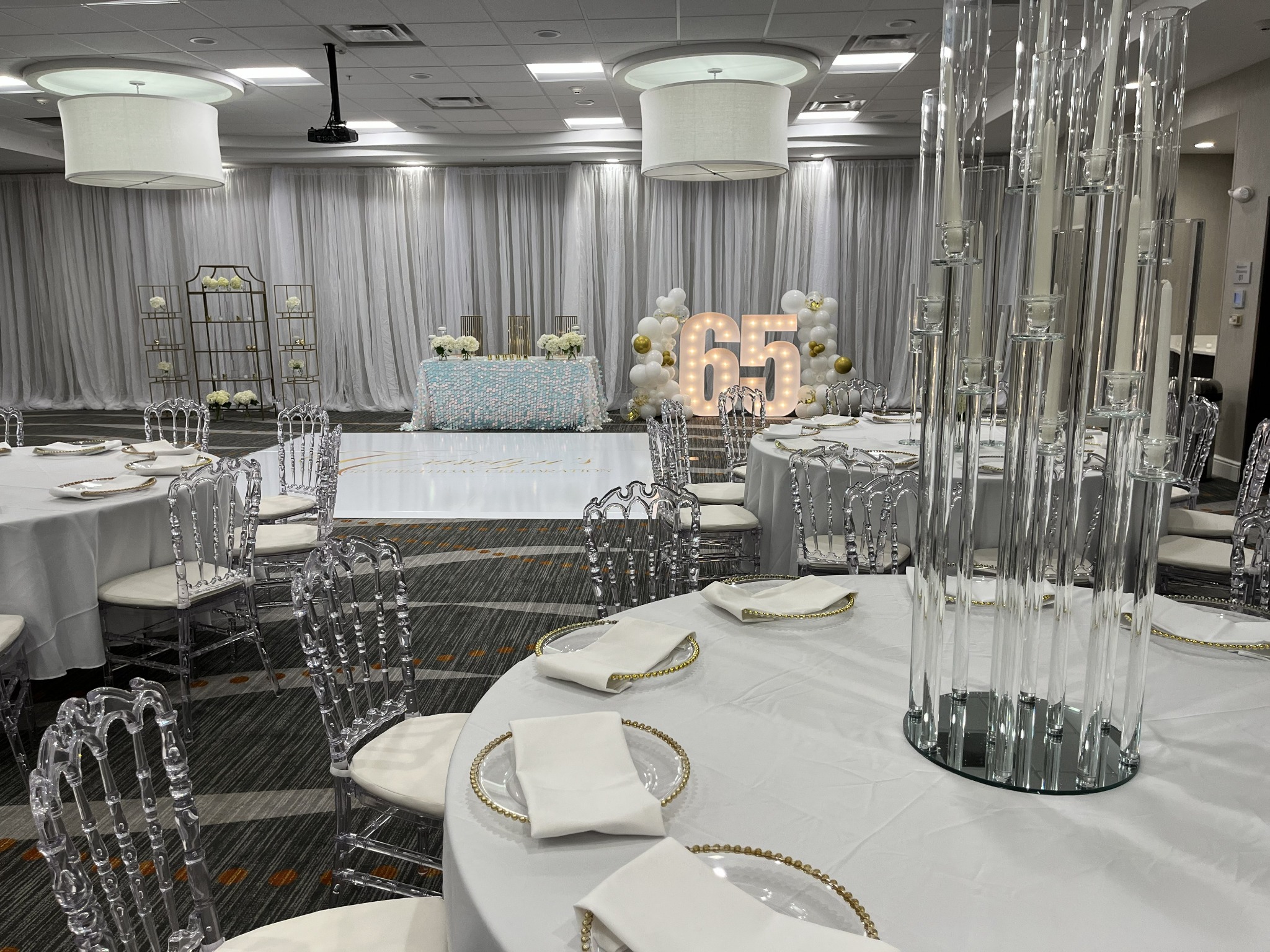 An all-white birthday room with clear chairs, tall glass candleholders, a personalized dance floor and illuminated number sixty-five