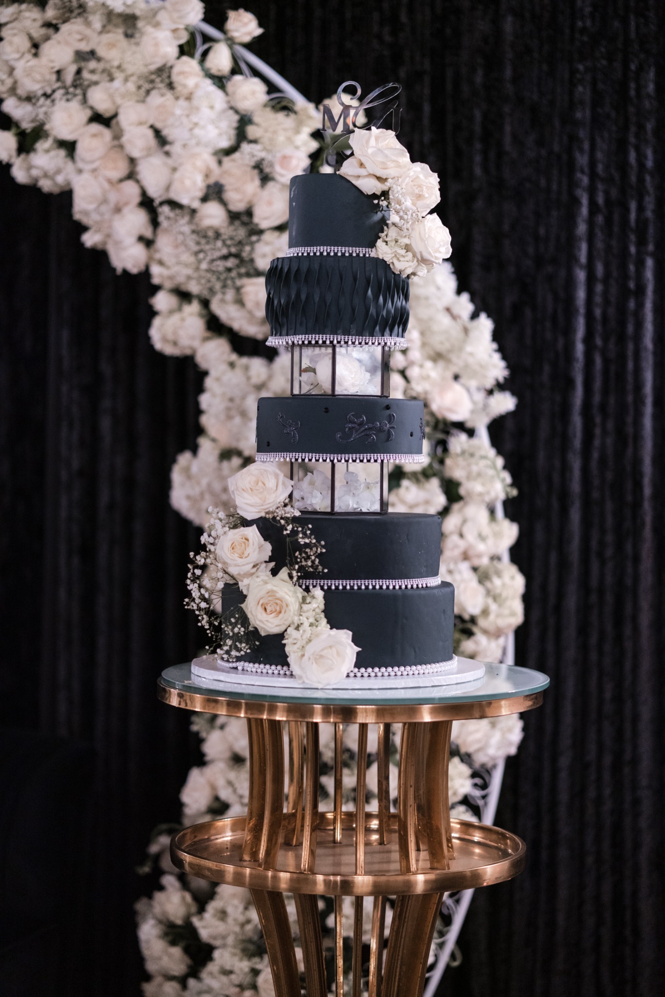 A sculptural black tiered cake with white roses and clear floral-filled tiers on a gold stand