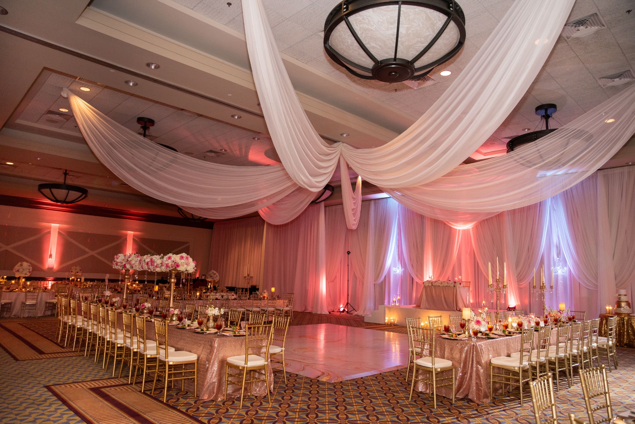 Sweeping ivory ceiling drapes, blush uplighting, gold chairs and floral tables surrounding a white dance floor