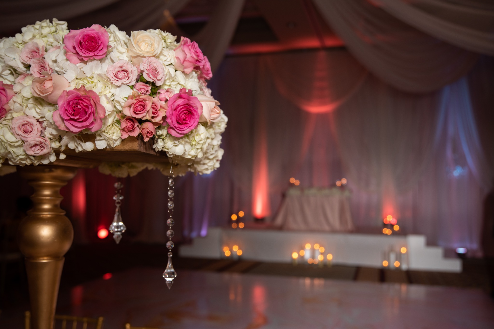 Pink and ivory roses on a gold pedestal with hanging crystals and softly lit draping