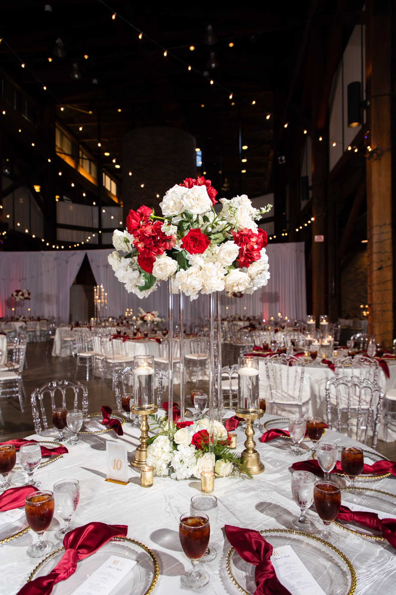 Red and ivory flowers on a tall clear stand above a white table with burgundy napkins, gold candleholders and glass place settings