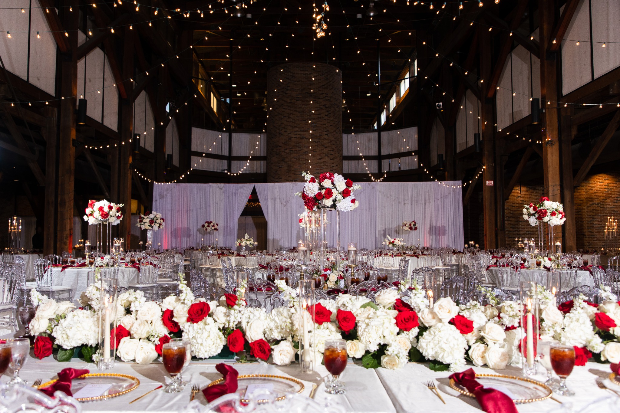 A reception room with red and ivory floral arrangements, clear chairs, white draping and warm overhead string lights